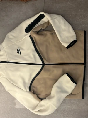 Nike tech fleece beige - Snygg hoodie från Nike i beige och vitt med svarta detaljer. Dragkedja framtill, huva och fickor på sidorna. Tillverkad i mjukt fleecematerial som är perfekt för en avslappnad och sportig stil.
