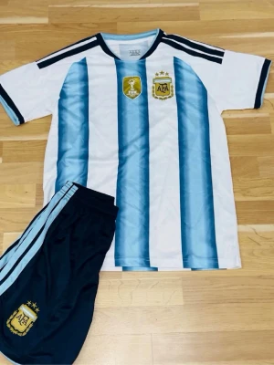 Argentina fotbollströja och shorts - Säljer ett matchande set med Argentina fotbollströja och shorts. Tröjan har klassiska blå och vita ränder, korta ärmar och tryckta emblem på bröstet. Shortsen är svarta med ljusblå ränder och emblem. Perfekt för fotbollsträning eller match.