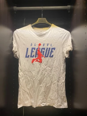 Vit t-shirt med Global League-tryck - Vit t-shirt med blå texten 'GLOBAL LEAGUE' och en röd siluett av en basketspelare på bröstet. Klassisk rund hals och korta ärmar. Perfekt för dig som gillar sportig stil och vill sticka ut med ett coolt tryck.