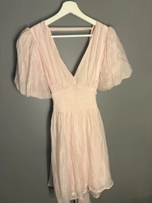 Ljusrosa kortklänning med puffärm - Söt ljusrosa kortklänning med djup v-ringning och voluminösa puffärmar. Klänningen har markerad midja och ett skirt, lätt material som ger ett romantiskt intryck. Perfekt för sommarens alla tillfällen.