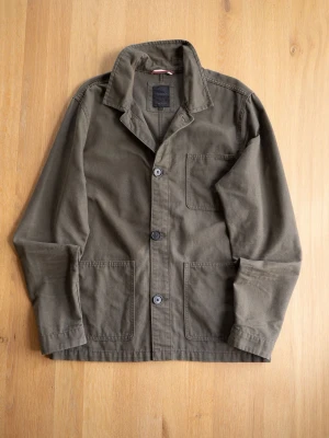 Olive Chore Jacket, size M - Stilren olivgrön overshirt i bomull med tre stora utanpåfickor och knäppning framtill. Klassisk krage och raka ärmar ger en avslappnad look. Perfekt att bära öppen eller stängd över t-shirt eller hoodie.