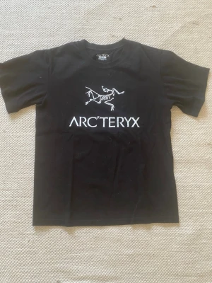 Svart Arc'teryx t-shirt med tryck - Snygg svart t-shirt från Arc'teryx med vitt logotryck och fossilmotiv på bröstet. Klassisk passform med rund hals och korta ärmar. Perfekt för dig som gillar outdoor-vibbar och stilren design. Tillverkad i mjuk bomull för skön känsla hela dagen. Liten i storlek