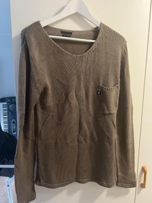 Brun stickad tröja med bröstficka - Säljer en brun stickad tröja med rund halsringning och en liten bröstficka på vänster sida. Tröjan har långa ärmar och en perfekt passform. Perfekt för lager på lager och enkel att matcha med olika stilar.