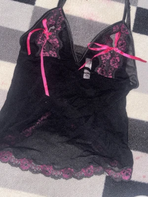 Svart spetslinne från Victoria's Secret - Säljer en svart mesh linne från Victoria's Secret med rosa spetsdetaljer och dekorativa rosetter framtill. Bodyn har tunna axelband och genomskinligt material med spetskant längst ner. Perfekt för dig som gillar feminina och lekfulla detaljer.
