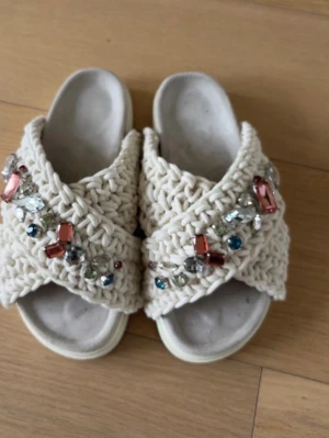 Inuikii - Unika inuikii- sandaler med breda, virkade band i vitt och stora dekorativa stenar i olika färger. Sandalerna har öppen tå och en bekväm sula, perfekta för dig som vill sticka ut med en bohemisk och trendig stil. Materialet är textil med inslag av plast och glas i dekorationerna.