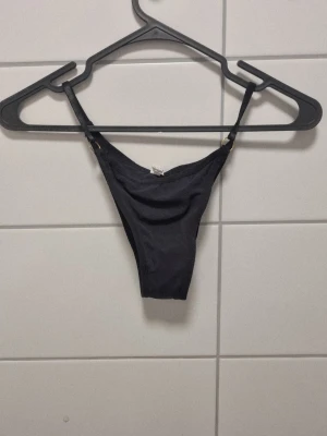 Svart trosa med ringdetaljer - Snygg svart trosa med tunna axelband och guldfärgade ringdetaljer på sidorna. Tillverkad i ett tunt, stretchigt material som känns mjukt mot huden. Perfekt för dig som gillar minimalistisk och stilren design.