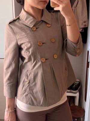 Kort beige trenchcoat med träknappar - Snygg kort trenchcoat i beige med dubbelknäppning och stora träknappar. Jackan har trekvartsärmar, bred krage och två fickor framtill. Tillverkad i ett mjukt bomullsliknande material som ger en avslappnad men stilren look. Gråbrun färg