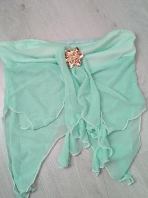 Mintgrön top med gulddetalj - Säljer en mintgrön top  i tunn, luftig syntet med en dekorativ guldfärgad blomma framtill. Modellen har asymmetriska kanter och ett skirt, draperat tyg som ger en unik look. Perfekt för stranddagar och sommarens soliga äventyr!