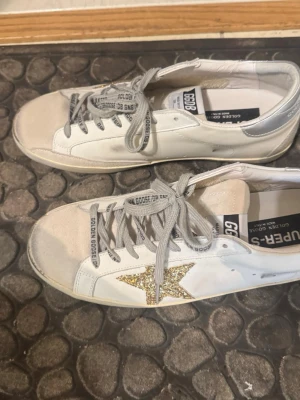 Vita Golden Goose sneakers med glitterstjärna - Säljer ett par vita Golden Goose sneakers i perfekt skick. Skorna har detaljer i mocka på tån och silverfärgad häl. Klassisk låg modell med text på snörningen och insidan. Ãkta kostar över 6000kr. Perfekt skick. Storlek 42-42.5.