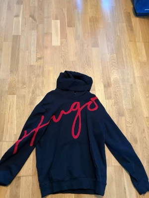 Hugo Boss Svart hoodie med röd text - Snygg svart hoodie med stor röd broderad text som går diagonalt över framsidan. Hoodien har huva och en klassisk magficka. Perfekt för dig som vill sticka ut med en unik detalj i vardagslooken.
