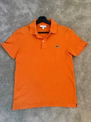Orange pikétröja från Lacoste - Klassisk orange pikétröja från Lacoste med korta ärmar och den ikoniska krokodilloggan på bröstet. Tröjan har krage och knäppning med två knappar framtill. Perfekt för en avslappnad och stilren look. Pikén är använd någon enstaka gång och säljes för den ej passar längre. 