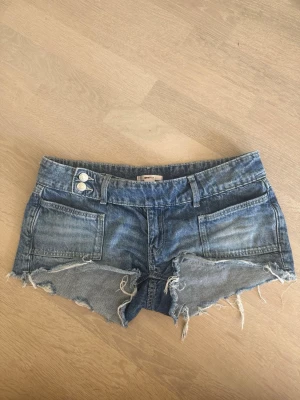 Blå jeansshorts - Säljer ett par blå jeansshorts med råa, fransiga kanter och slitningar. Shortsen har två stora fickor fram, två bakfickor och dubbla knappar i midjan. Perfekta för varma dagar och en avslappnad stil. Pris kan diskuteras!