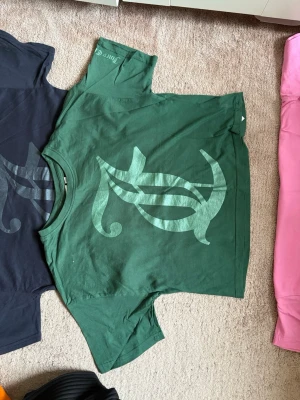 Grön t-shirt med tryck - Snygg grön t-shirt med ett stort grafiskt tryck på framsidan. T-shirten har korta ärmar och en klassisk rund halsringning. Perfekt för dig som vill ha en unik och stilren look.