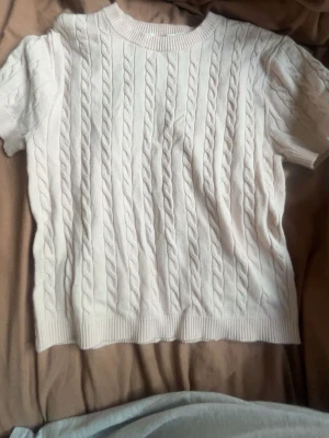Vit kabelstickad kortärmad tröja H&M - Snygg vit kabelstickad tröja från H&M med korta ärmar och rund halsringning. Tröjan har ribbade muddar vid hals, ärmslut och nederkant. Perfekt för en stilren och fräsch look till vardags.