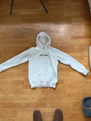 Vit hoodie från Palm Angels - Säljer en vit hoodie från Palm Angels med svart logga på bröstet och stor text på ryggen. Klassisk känguruficka framtill och ribbade muddar vid ärmar och nederkant. Perfekt för dig som gillar streetwear och vill ha en clean look.
