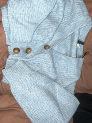 Ljusblå stickad tröja från H&M - Mysig ljusblå stickad tröja från H&M med stora bruna knappar vid axeln. Tröjan har ribbade muddar och en avslappnad passform. Perfekt för kalla dagar och enkel att styla med jeans eller kjol.