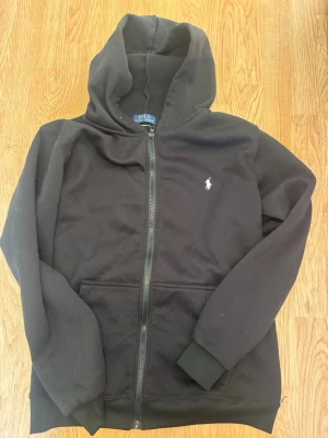 Svart hoodie från Polo Ralph Lauren - Klassisk svart hoodie med dragkedja från Polo Ralph Lauren. Hoodien har huva, fickor framtill och den ikoniska vita logobrodyr på bröstet. Tillverkad i mjukt material som passar perfekt för en avslappnad stil.