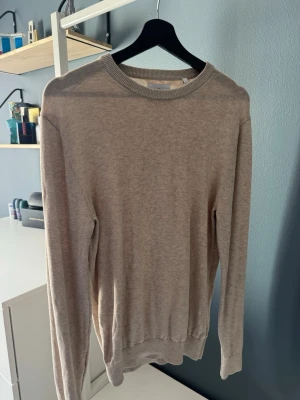 Beige stickad tröja med rund hals - En stilren beige stickad tröja med rund halsringning och långa ärmar. Tröjan har ribbade muddar vid ärmslut och nederkant. 