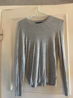 Ljusgrå stickad tröja - En enkel och stilren ljusgrå stickad tröja med rund halsringning och långa ärmar. Tröjan har ribbade muddar vid ärmslut och nederkant, vilket ger en klassisk look.