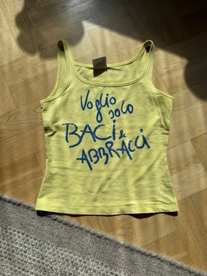 Gul linnetopp från Baci & Abbracci - Gul linnetopp från Baci & Abbracci med blå texttryck framtill. Toppen har smala axelband och är tillverkad i mjuk bomull, perfekt för varma dagar. Snygg och enkel att matcha med jeans eller kjol. Inga defekter och den är i strl S. Skriv vid frågor!🫰🏻