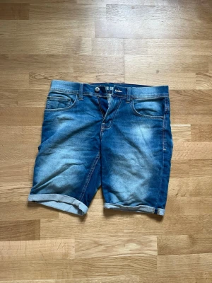 Blå jeansshorts med uppvik - Säljer ett par klassiska blå jeansshorts med uppvikta benslut. Shortsen har fem fickor, bälteshällor och stängs med knapp och dragkedja. Perfekta för sommaren och har en snygg tvättad look.