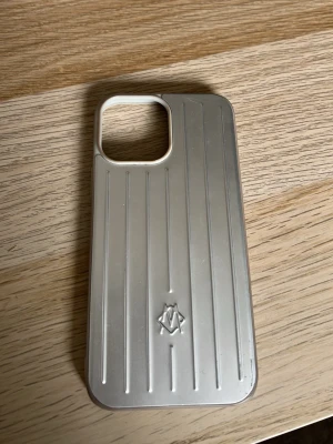 Silver mobilskal från Rimowa - Snyggt silverfärgat mobilskal från Rimowa med klassisk räfflad design och diskret logga på baksidan. Skalet är gjort i hårdplast och har en vit ram runt kamerauttaget. Passar perfekt för dig som gillar stilren och exklusiv look som passar till iPhone 14 Pro