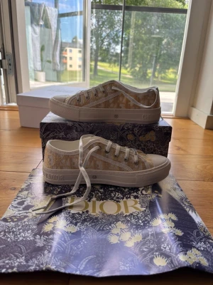 Walk’n dior sneakers ny - Säljer ett par snygga beige och vita Dior sneakers med klassiskt monogrammönster. Skorna har vita snören, gummisula och detaljer med Dior-logga på sidan och tungan. Perfekta för dig som vill ha en exklusiv touch till din outfit.