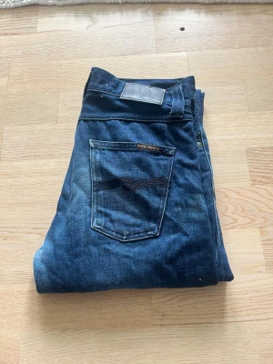 Mörkblå jeans från Nudie Jeans - Säljer ett par klassiska mörkblå jeans från Nudie Jeans. Jeansen har fem fickor, raka ben och snygga kontrastsömmar. Perfekta för en avslappnad och stilren look. Materialet är bomull och passformen är normal. W32 L32