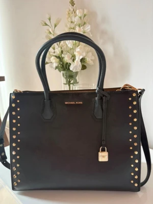 Svart handväska Michael Kors nitar - Snygg svart handväska från Michael Kors i slätt läder med guldfärgade nitar längs kanterna och ett dekorativt hänglås framtill. Väskan har både handtag och axelrem. 