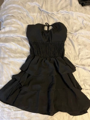 Svart kort volangklänning - Säljer en svart kort klänning med volanger och smock i midjan. Klänningen har axelbandslös design med knyt i nacken och flera lager i kjolen för en snygg look. Perfekt för dig som vill ha något stilrent och trendigt.