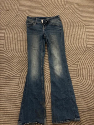 Blå bootcut jeans med slitningar - Säljer ett par klassiska blå bootcut jeans med lätt slitna detaljer framtill. Jeansen har normalhög midja och är tillverkade i denim. Perfekta för dig som gillar en avslappnad och tidlös stil. De är också slitna längst ner till byxan men är fortfarande i väldigt bra skick 