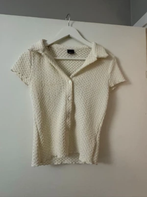 Vit stickad blus med krage - Snygg vit stickad blus med korta ärmar och klassisk krage. Blusen har knäppning framtill med vita knappar och ett strukturerat, bubbligt mönster i materialet. Perfekt för en trendig och avslappnad look.