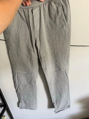 Grå linnebyxor med resårmidja - Snygga grå linnebyxor med avslappnad passform och resår i midjan. Byxorna har sidfickor fram och två bakfickor med knapp. Perfekta för en chill och stilren look. Materialet är lätt och luftigt, vilket gör dem sköna att bära under varma dagar.
