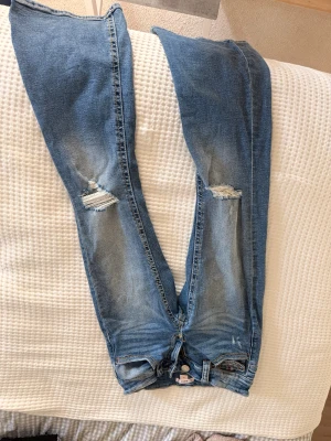Blå slitna jeans från Gina Tricot - Säljer ett par blå jeans från Gina Tricot i storlek 158. Jeansen har skinny fit, slitningar på båda knäna och en klassisk femficksmodell. Materialet är denim med stretch för extra komfort. Perfekta för en avslappnad och trendig look.