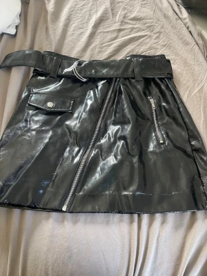 Svart lackkjol med bälte och dragkedja - Snygg svart kortkjol i glansigt lackmaterial med bälte i midjan. Kjolen har en sned dragkedja framtill, en ficka med knapp och en extra dragkedjedetalj. 