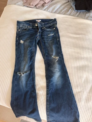 Blå slitna jeans från Gina Tricot - Ett par blå jeans från Gina Tricot med slitningar och hål på benen för en trendig look. Jeansen har klassisk femficksmodell, låg midja och utsvängda ben. Tillverkade i denim med vita kontrastsömmar och dragkedjegylf.