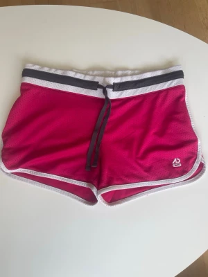 Rosa sportiga nike shorts med snörning - Snygga rosa sportshorts med vita och grå detaljer i midjan samt vit kant runt benen. Shortsen har snörning i midjan och är tillverkade i ett lätt och ventilerande syntetmaterial, perfekta för träning eller varma dagar. Storlek:S-M