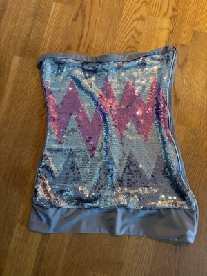 Glittrig tubtopp med paljetter - Säljer en glittrig tubtopp med paljetter i silver och rosa, dekorerad med ett zigzag-mönster. Toppen är axelbandslös och har en bandeauformad passform med breda kanter upptill och nedtill i grått tyg. Perfekt för dig som vill sticka ut.