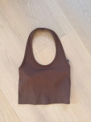 Brun ribbad linnetopp - Säljer en brun ribbad linnetopp med rund halsringning och ärmlös design. Toppen är croppad och har en enkel, stilren look som passar perfekt till jeans eller kjol. Materialet känns mjukt och stretchigt för en bekväm passform.