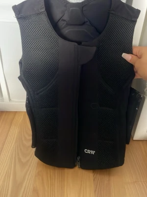 Svart skyddsväst ryggplatta CRW - Säljer en svart skyddsväst från CRW med meshpaneler för extra ventilation och vaddering för bra skydd. Västen har dragkedja framtill och reflekterande detaljer på ryggen. Perfekt för ridning. Skyddspadda baktill men väldigt tunn annars, så funkar perfekt under en jacka eller tjocktröja.