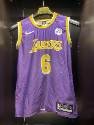  LeBron James jersey #6 - Snyggt basketlinne från Nike med Lakers tryck och nummer 6 för LeBron James. Linnet är lila med gula och vita detaljer, ärmlöst och har officiella NBA-märken. Perfekt för basketträning eller att bära som supporter.