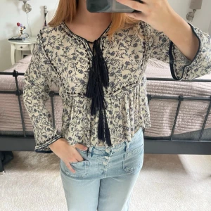Blommig boho-blus med knytband - Supersöt boho-inspirerad blus i beige med mörkblått blommönster. Blusen har långa ärmar, volang nedtill och dekorativa knytband med tofsar framtill. Materialet känns lätt och luftigt, perfekt för en avslappnad och trendig look.
