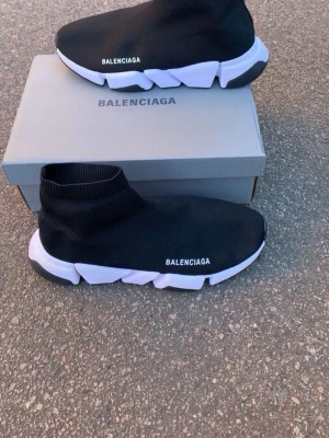  Speed Trainer svarta sneakers - Speed Trainer sneakers med vit chunky sula och stickad ovandel i textil. Strumpmodell med logga på sidan och unik, futuristisk sula i svart och vitt. Perfekta för dig som gillar streetwear och vill ha något riktigt trendigt.
