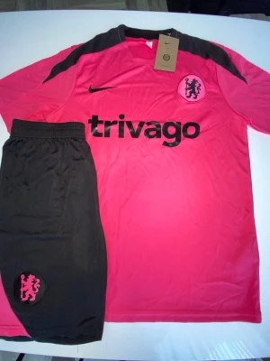 Chelsea rosa träningsset Nike - Säljer ett rosa och svart Chelsea träningsset från Nike. Setet består av en kortärmad tröja med svart logga och tryck samt matchande svarta shorts med rosa Chelsea-märke. Materialet är lätt och andas, perfekt för fotbollsträning eller gym.