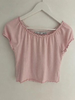 Ljusrosa croppad topp med detaljer - Söt ljusrosa croppad topp med volangkant vid ärmslut, halsringning och nederkant. Toppen har korta ärmar och en lätt rynkad struktur som ger en avslappnad och trendig look. Perfekt för varma dagar och enkel att styla. Ganska liten i storlek.