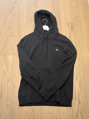 J Lindeberg hoodie  - Snygg svart hoodie från j lindeberg med huva och dragsnören. Har den vanliga j lindeberg loggan på bröstet och en klassisk känguruficka framtill. Perfekt för en avslappnad och stilren look.