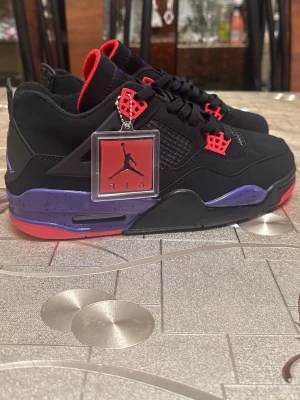 Svarta Air Jordan 4 Retro sneakers - Säljer ett par Air Jordan 4 Retro sneakers i svart med röda och lila detaljer. Skorna har meshpaneler, röda snörningsdetaljer och en specklad lila mellansula. Klassisk Jumpman-tag på sidan och broderad logga på plösen. Perfekta för dig som gillar streetwear.