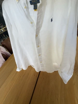 Vit linneskjorta från Polo Ralph Lauren - Klassisk vit skjorta i linne från Polo Ralph Lauren med knappslå och broderad logga på bröstet. Skjortan har lång ärm, button-down krage och en avslappnad passform. Perfekt för en stilren och fräsch look. Ej använd. Tagit bort lappen. Priset ej hugget i sten. Dm för frågor 