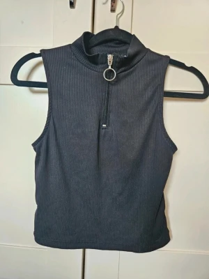 Topp lager 157 M L - Svart croppad ärmlös topp från 157 med ribbad struktur och hög krage. Toppen har en snygg dragkedja framtill med rund metallring som detalj. Perfekt att styla med jeans eller kjol för en cool look.  Kan även hämtas I Partille  E1