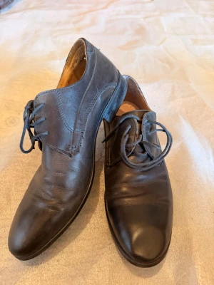 Svarta klassiska läderskor med snörning - Ett par svarta klassiska läderskor med rundad tå och snörning. Skorna har låg klack och stilrena sömmar längs sidorna. Insidan är ljusbrun och materialet är slätt läder. Perfekta för dig som gillar enkel och tidlös design.